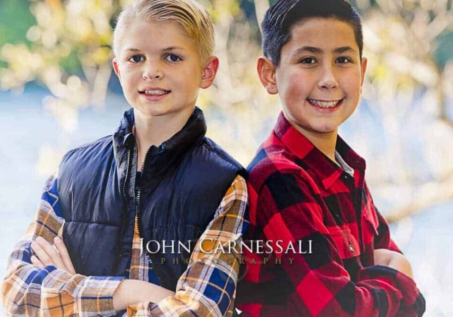 Two boys smiling outdoors, one in a blue vest and the other in a red plaid shirt.
