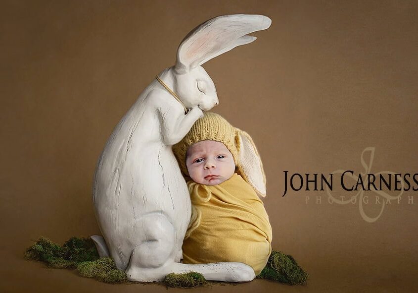 Baby dressed as a carrot next to a large white rabbit statue.