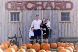 John Carnessali Photography Family of four posing with pumpkins in front of a rustic wooden building.