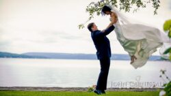 Groom lifting bride by lakeside under tree.